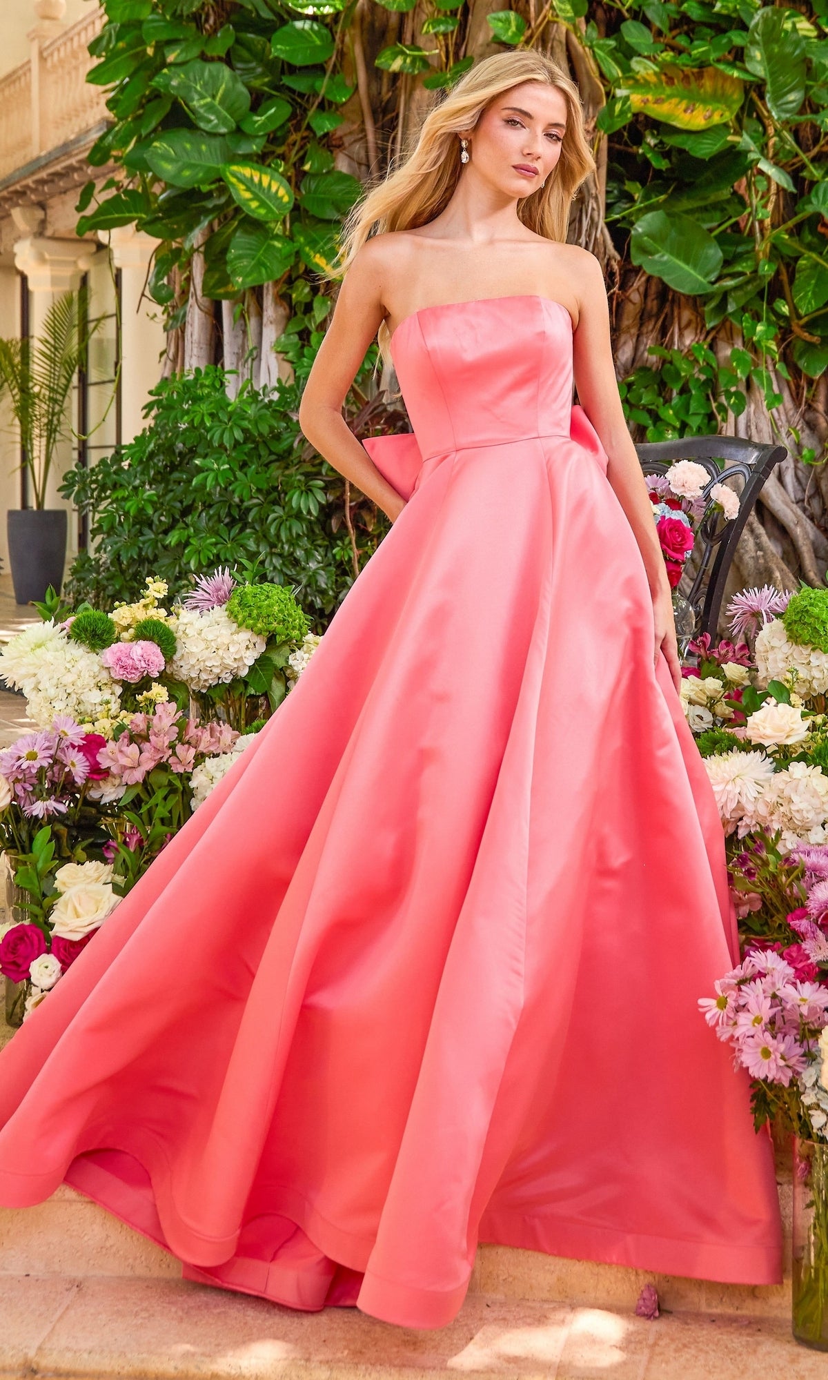 A woman wearing a strapless, floor-length pink satin Ava Presley 42523 ball gown with a large bow on the back, standing among flowers.