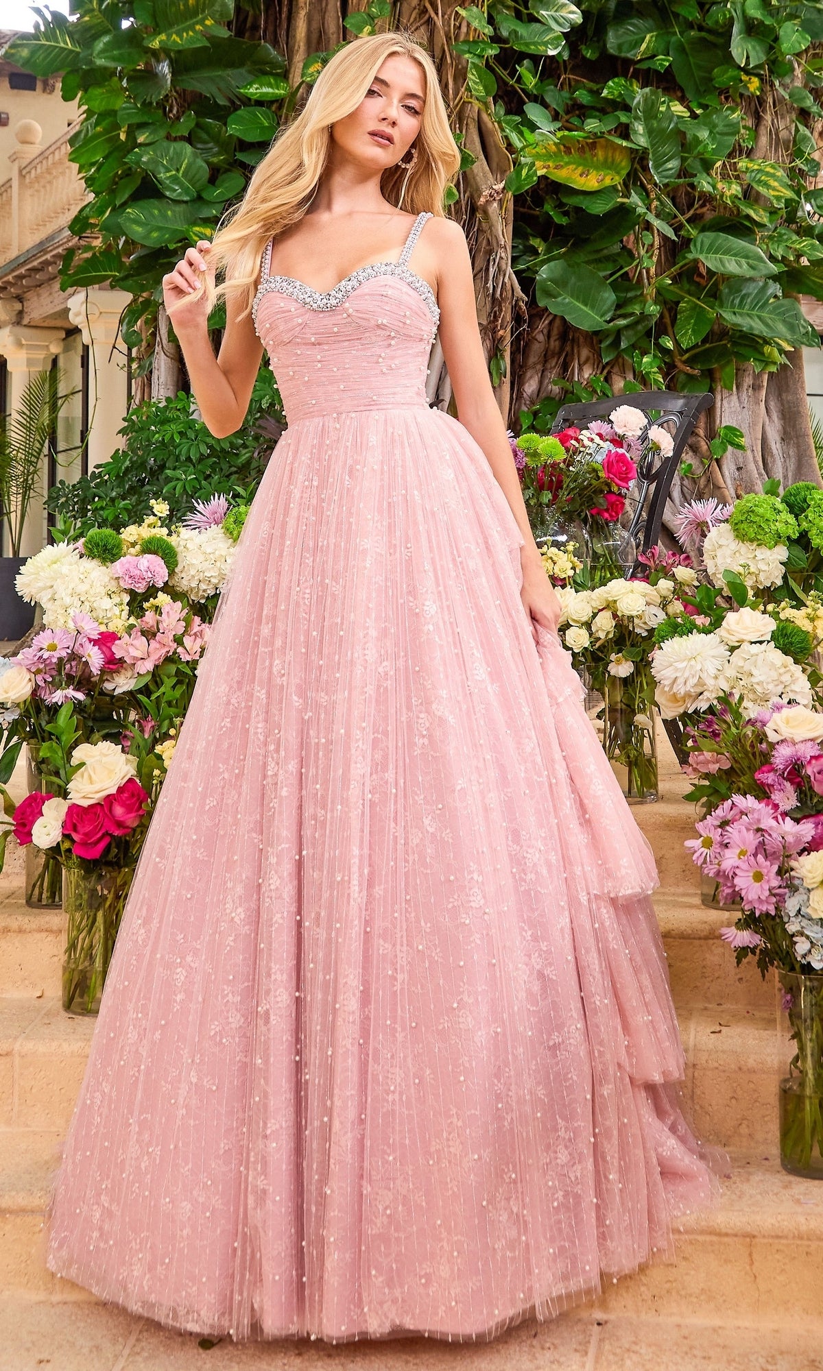 A woman wearing a floor-length, beaded pink sweetheart Ava Presley 42526 prom dress with rhinestone-trimmed straps, standing among vases of flowers.