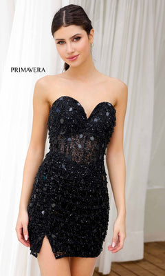 A woman stands before white curtains wearing the Beaded Strapless Short Hoco Dress: Primavera 4252, a black sequin-embellished mini with a front slit.