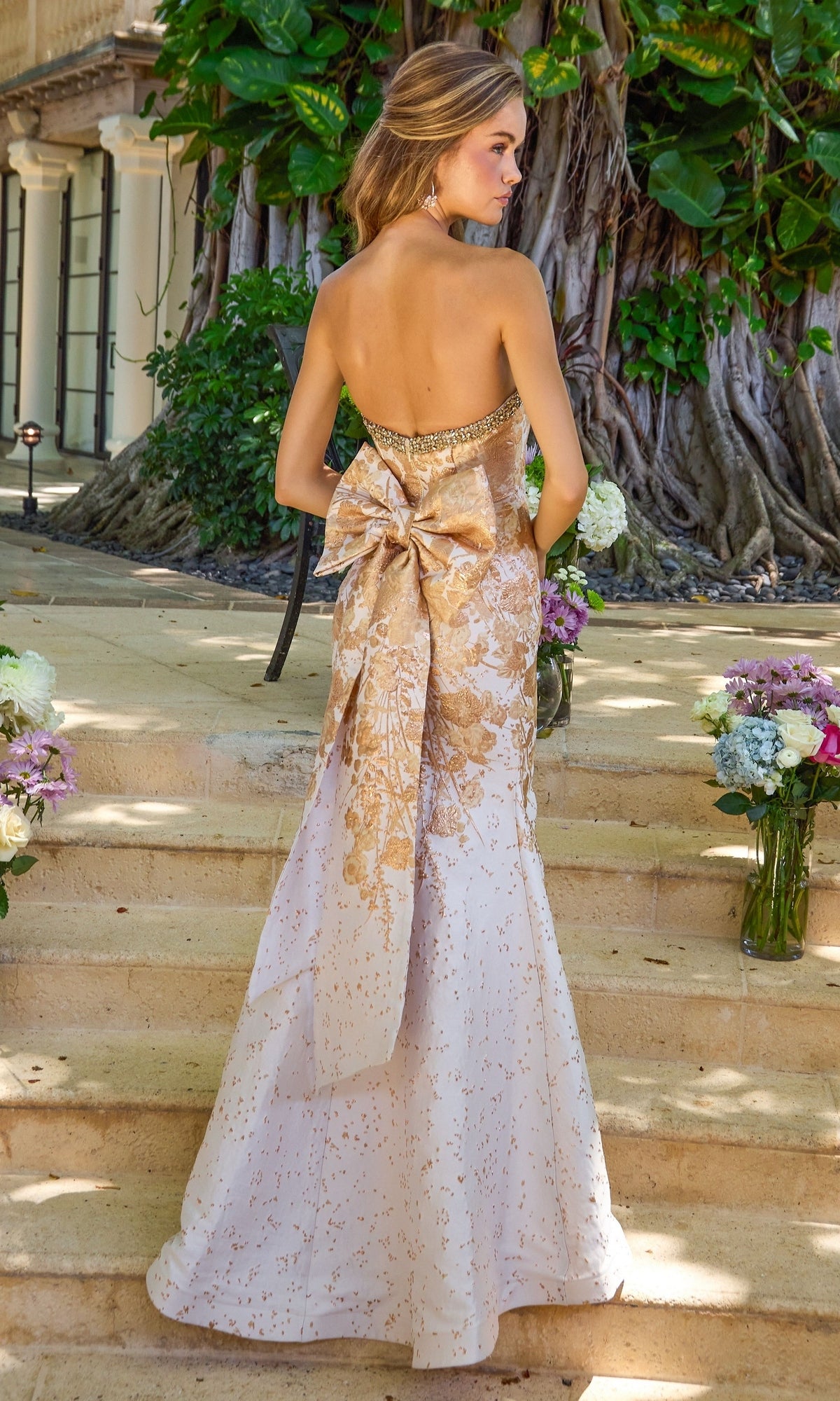 Long Prom Dress: Ava Presley 42535, a strapless white floor-length mermaid gown featuring a metallic gold floral pattern and a large bow at the lower back.