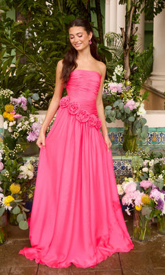 A model wears a bright pink Long Prom Dress: Ava Presley 42536, a strapless floor-length gown with gathered fabric and a rosette cluster at the hip, standing in front of floral arrangements.