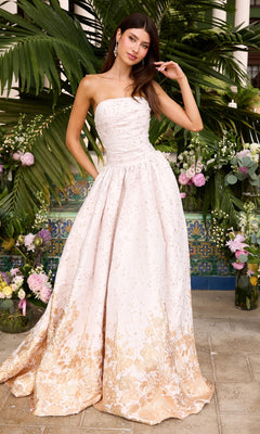 A person wears an Ava Presley 42540 strapless, floor-length light pink floral prom gown with a gold hem pattern while standing in front of tropical plants and flowers.