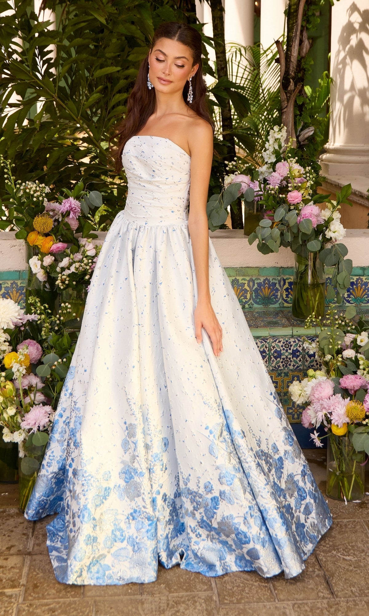 A woman wears an Ava Presley 42540 strapless, floor-length gown with a white bodice and a blue floral pattern that fades toward the hem while standing in front of flower arrangements.