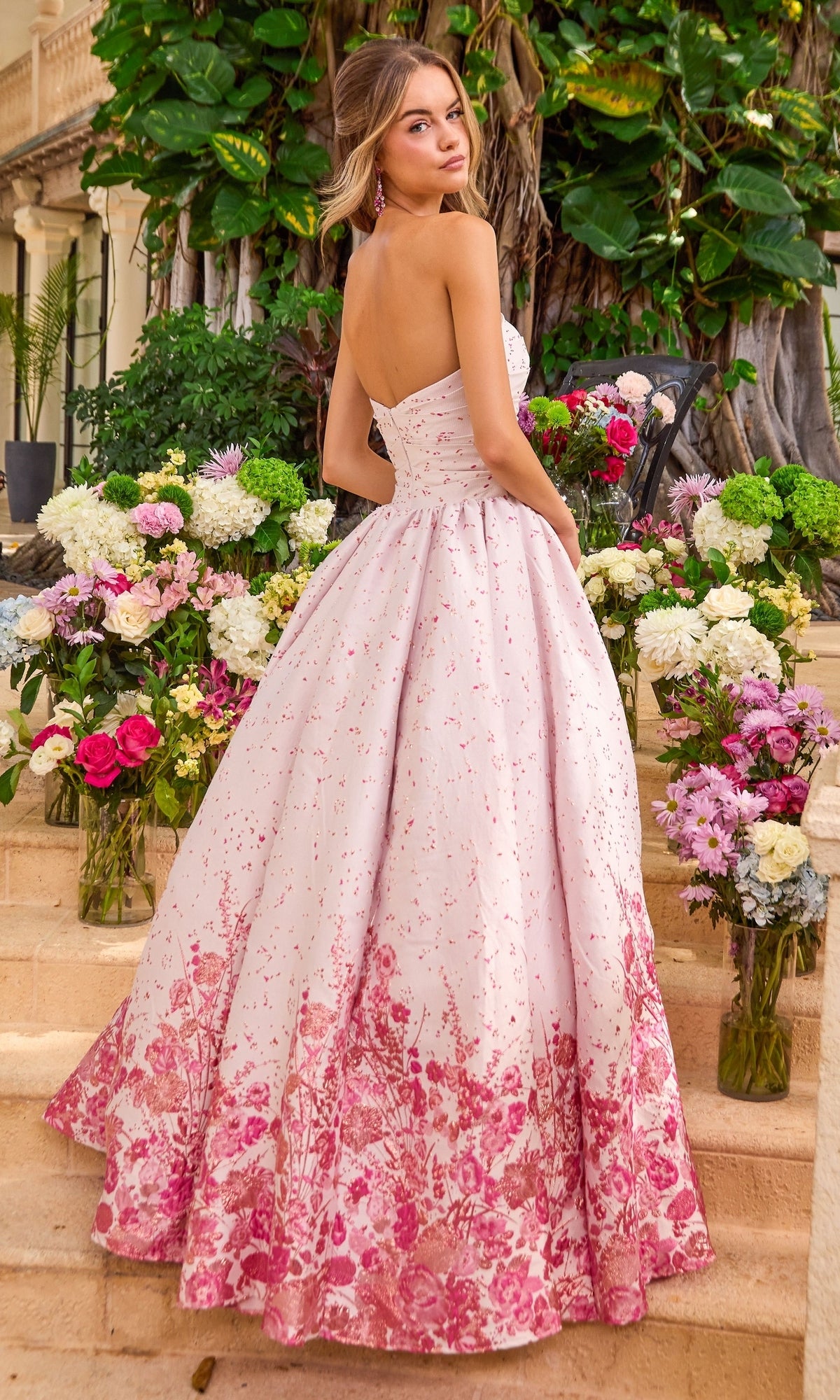 A person wearing a light pink Ava Presley 42540 strapless ballgown with a dark pink floral print on the skirt, standing on stone steps surrounded by flower arrangements.