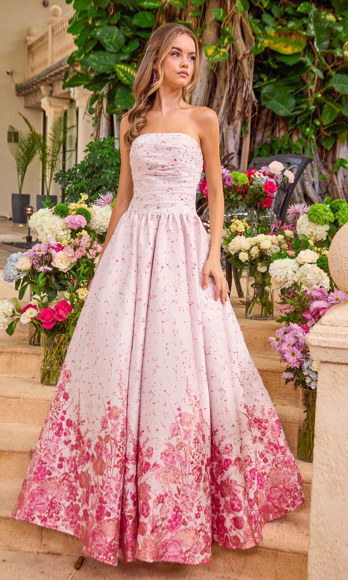 A woman wearing an Ava Presley 42540 strapless floral prom ball gown with a pink petal print and a dark floral pattern along the hem, standing among flower arrangements on stone steps.