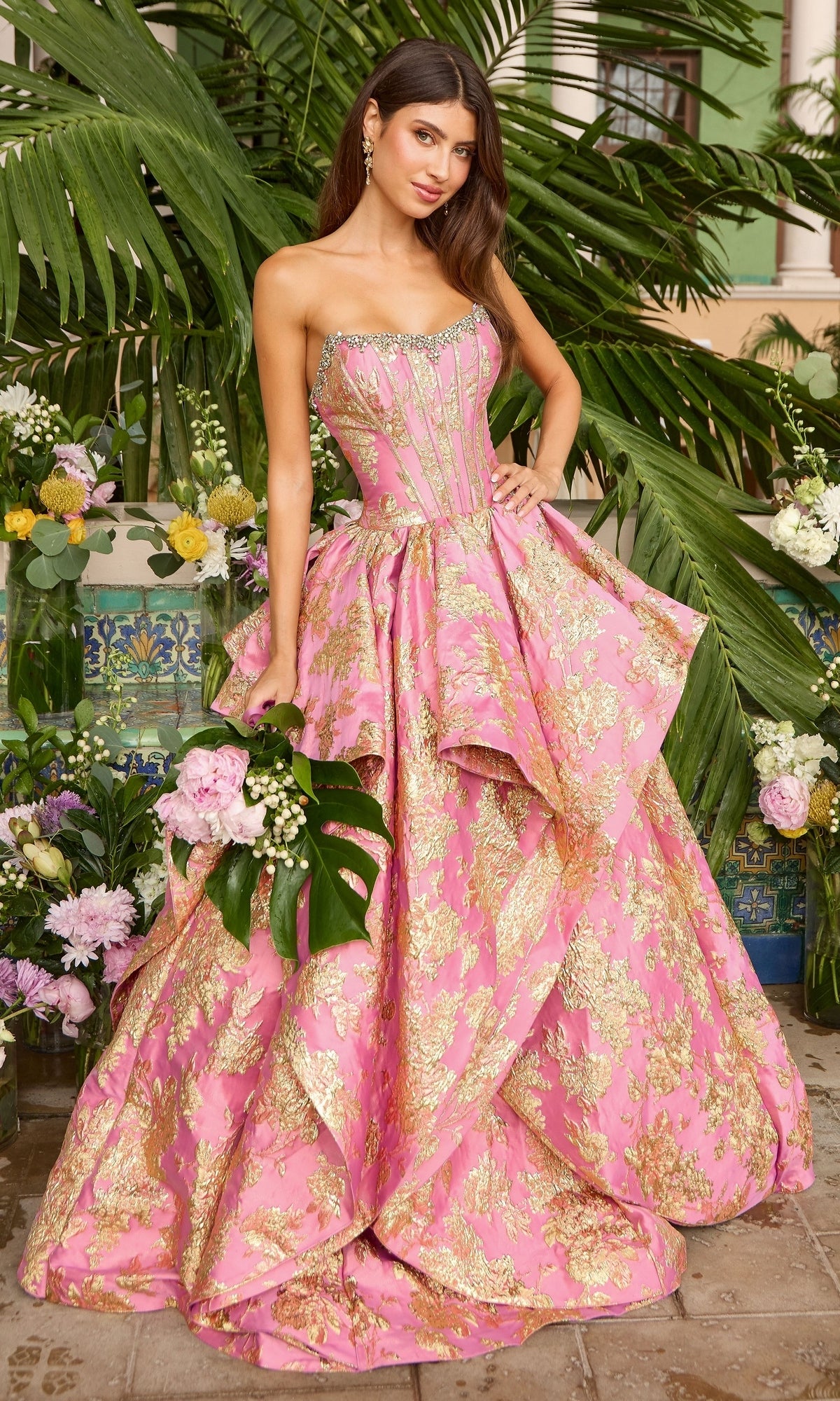 A woman wears a strapless pink and gold Ava Presley 42541 brocade gown with a corset bodice and a full skirt, holding a bouquet of flowers.