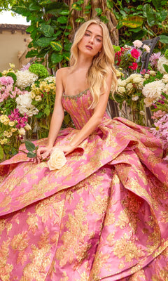 A person sits among potted flowers wearing a strapless, pink and gold metallic Ava Presley 42541 ballgown with a ruffled skirt.