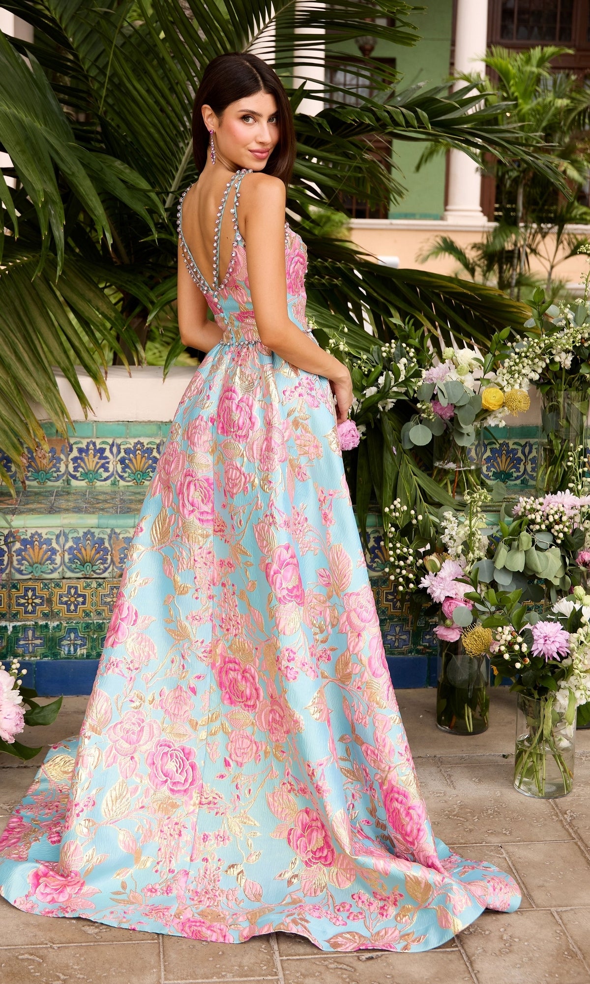 A woman wears an Ava Presley 42549 floral A-line prom dress featuring a floor-length sleeveless gown with a blue, pink, and gold brocade print and beaded straps.