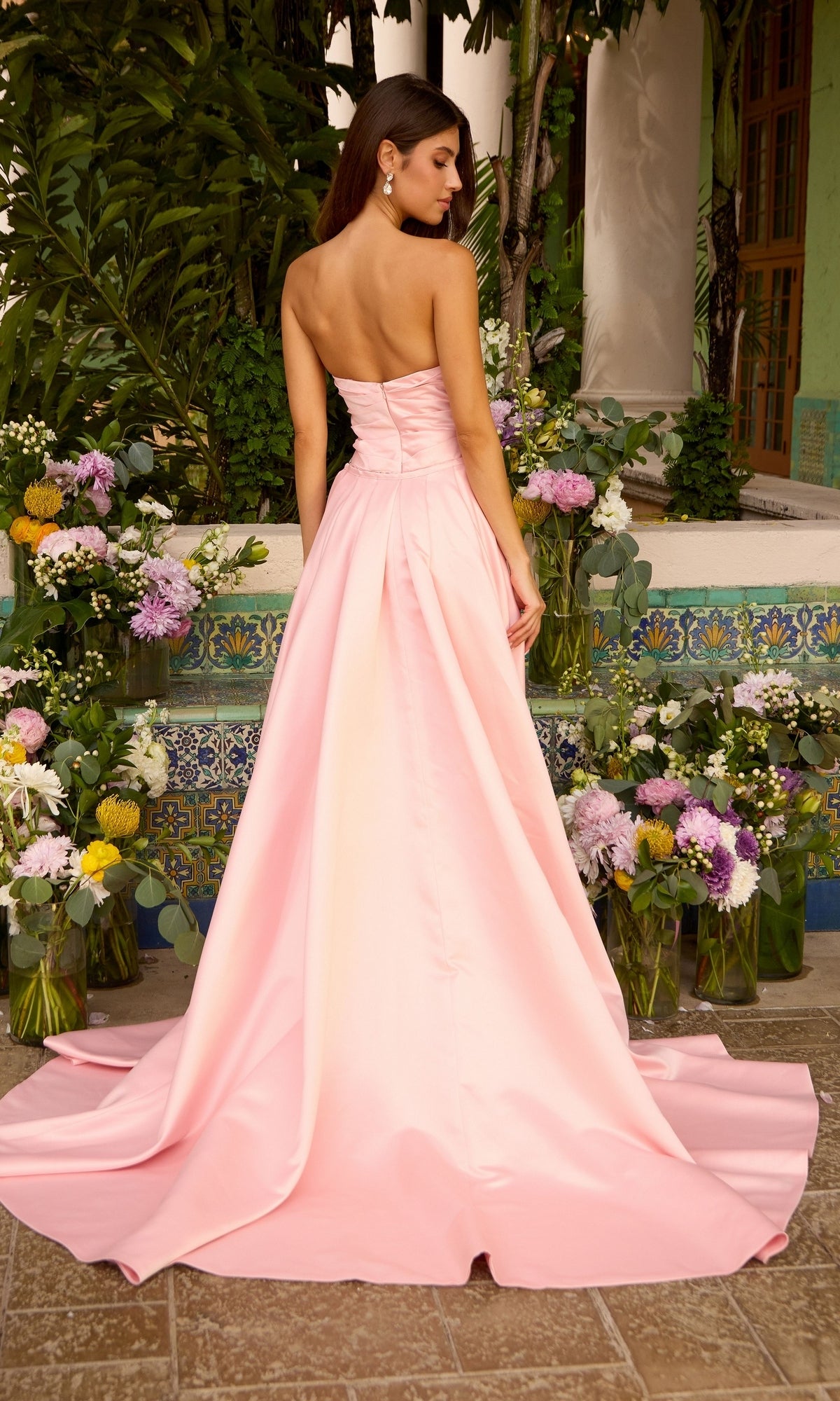 Back view of a light pink Ava Presley 42553 fitted prom dress with a full overskirt and floor-length train, displayed in front of floral arrangements.