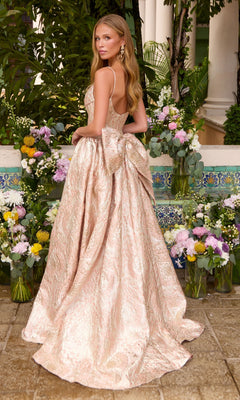 A person wearing an Ava Presley 42554 shimmering, floor-length pale pink metallic floral ball gown with a large waist bow, standing in front of floral arrangements.