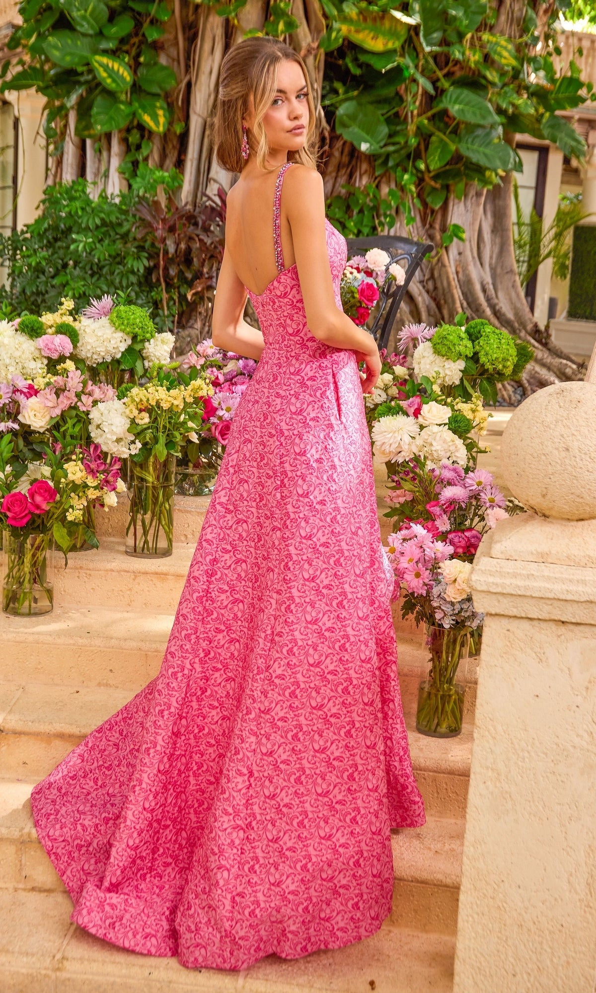 A woman stands on stone steps surrounded by flowers wearing a floor-length, backless Ava Presley 42577 A-line prom dress with a repeating floral pattern.