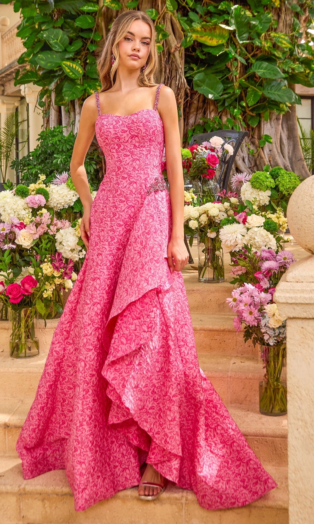 A hot pink patterned Ava Presley 42577 A-line prom dress with beaded straps and a side-slit skirt, standing amidst floral arrangements.