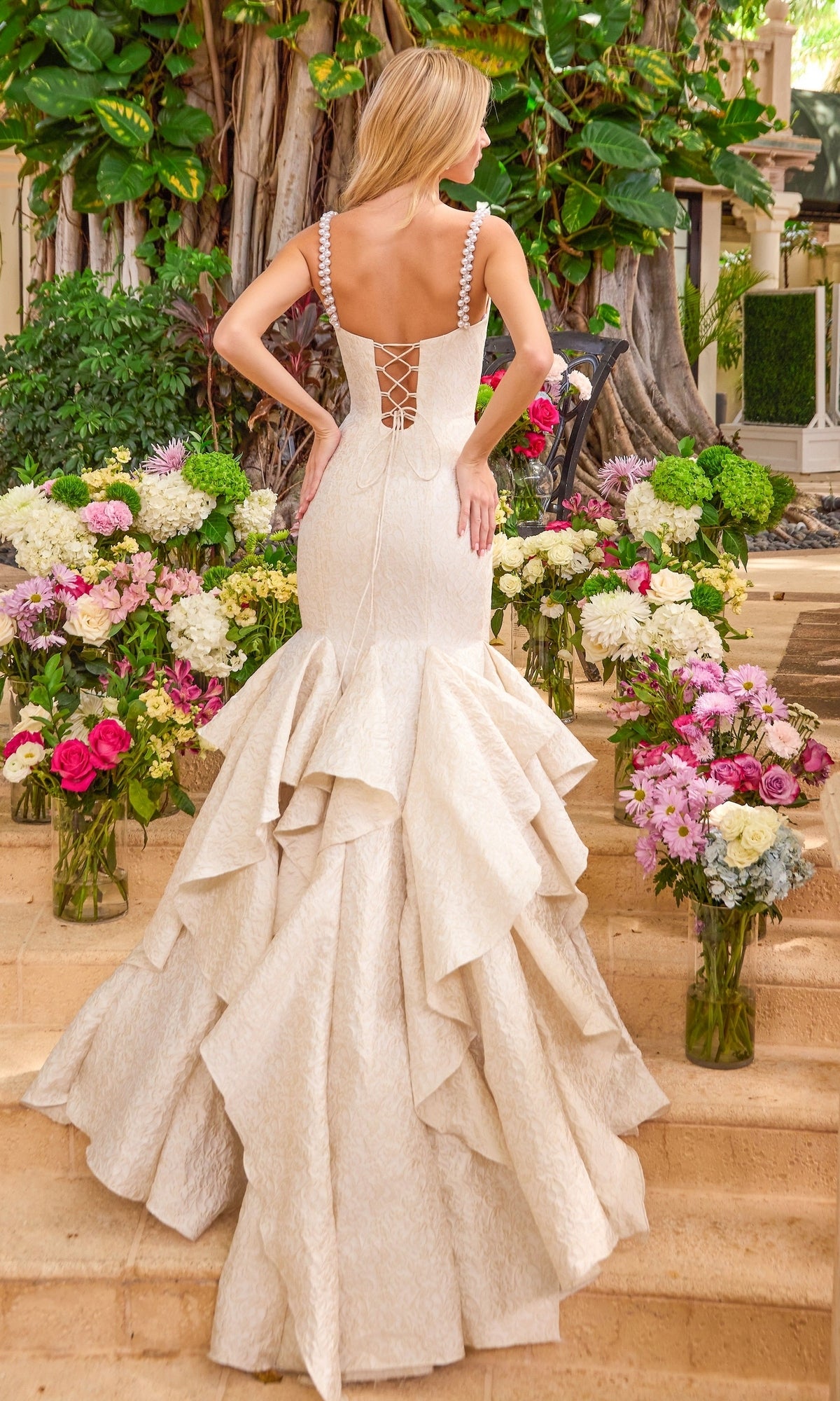 Back view of an off-white Long Prom Dress: Ava Presley 42578 featuring a lace-up back and ruffled floor-length skirt, worn by a woman standing on steps among vases of flowers.