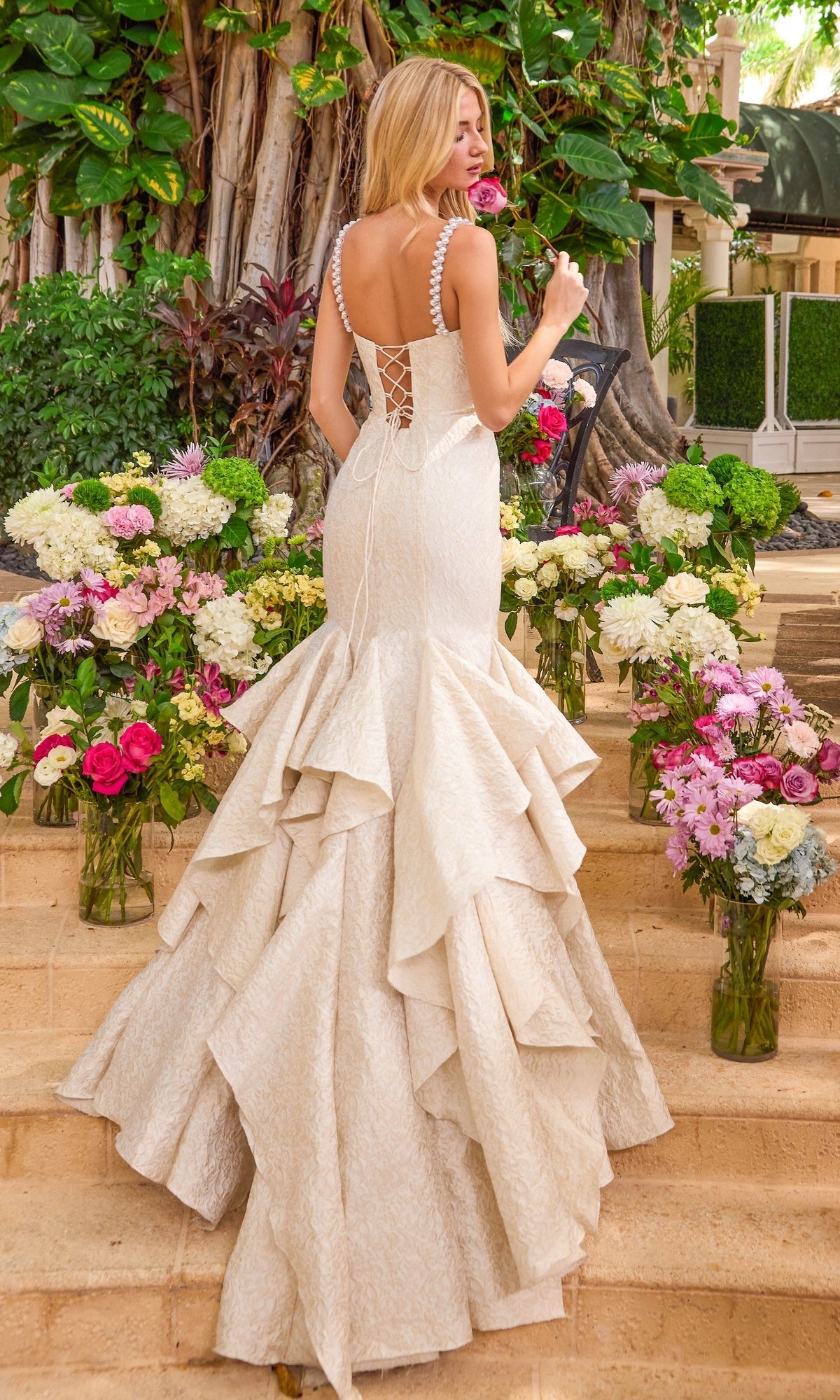 A woman wears a white Ava Presley 42578 gown with pearl-trimmed straps, a lace-up back, and a tiered ruffled skirt while standing among vases of flowers on stone steps.