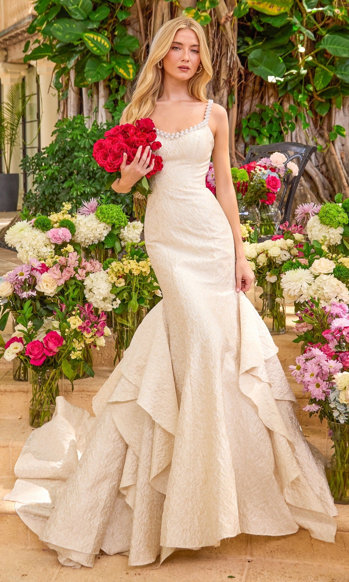 A woman in a cream-colored, textured Ava Presley 42578 mermaid prom dress with pearl-embellished straps holds a bouquet of red roses, surrounded by various floral arrangements.