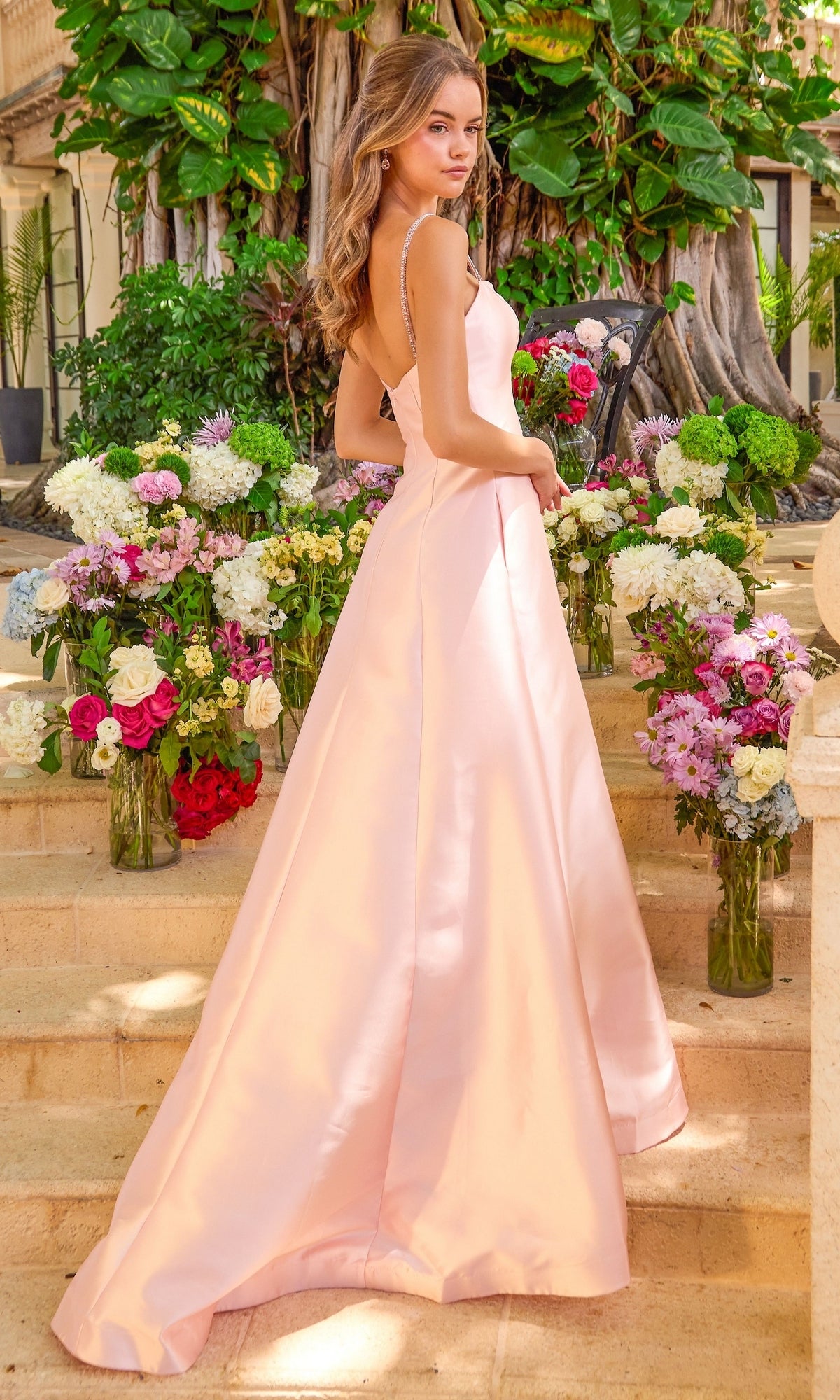 A woman stands on stone steps surrounded by vases of flowers wearing a light pink, floor-length, ruffle A-line Ava Presley 42579 prom dress with spaghetti straps.