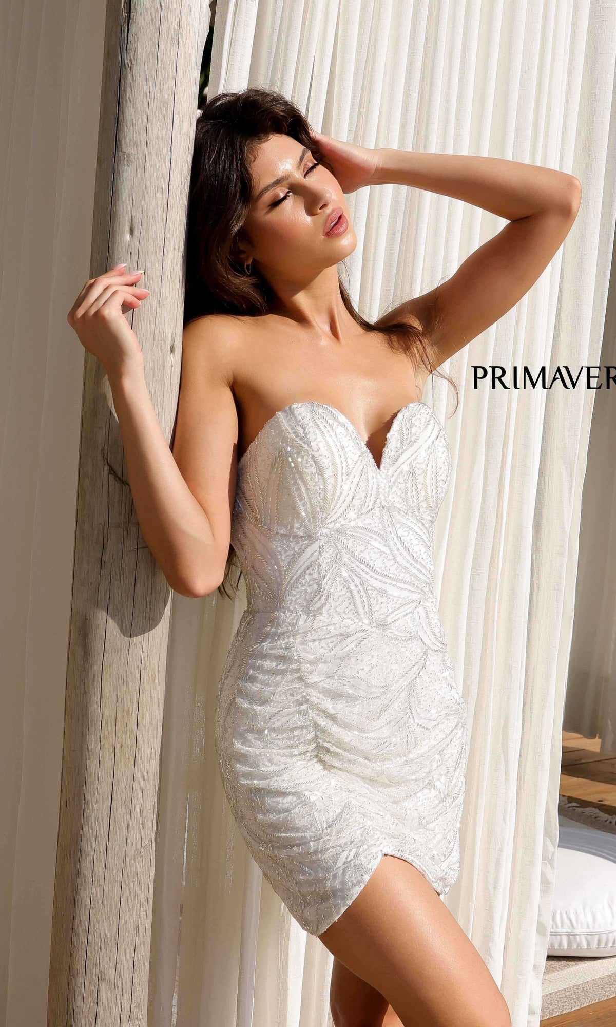 A woman poses in front of white curtains, one hand on her head, wearing the Primavera 4259 Strapless Beaded Mini Hoco Dress with a sweetheart neckline.