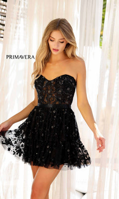 A woman in the Short Homecoming Dress 4263 by Primavera, featuring a strapless sweetheart neckline, black lace, floral embroidery, fitted waist, and sheer bodice, stands near white curtains in this chic party look.