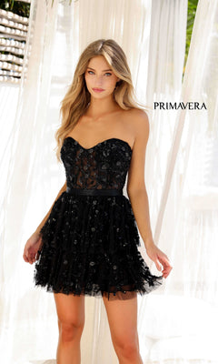 A woman wears the Short Homecoming Dress 4263 by Primavera, featuring a strapless sweetheart neckline, black lace, and a tiered skirt, while standing before sheer white curtains with "PRIMAVERA" visible in the background.