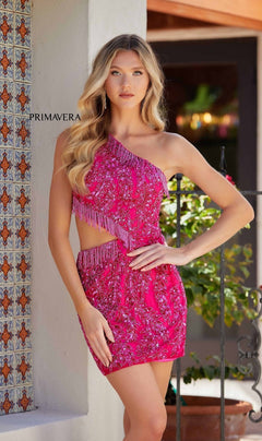 A woman poses outdoors in the Beaded Short Cut-Out Hoco Dress: Primavera 4264—a bright pink, one-shoulder dress with sequins and fringe. A "Primavera" sign is visible on the wall behind her.