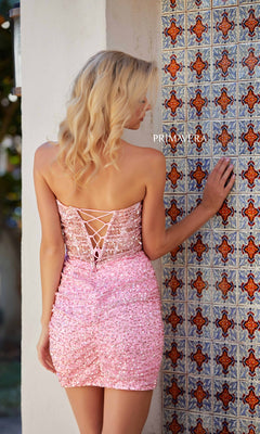 A woman with blonde hair wears the Short Homecoming Dress 4266 by Primavera—a sparkly pink, strapless sequin dress with a lace-up back—standing by a patterned tiled wall and facing away from the camera.