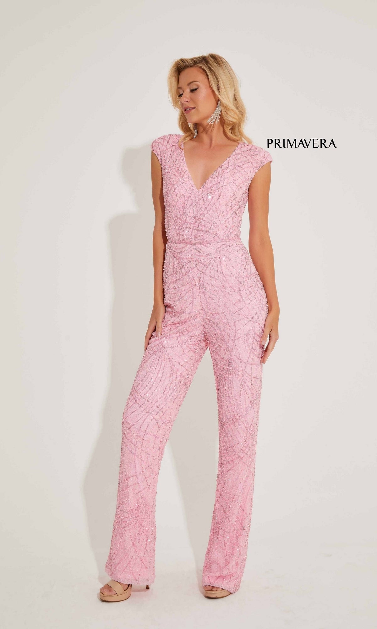 A woman stands against a white background wearing the Long Formal Jumpsuit 4275 by Primavera—a pink, sleeveless, V-neck jumpsuit with sequin detailing. The word 
