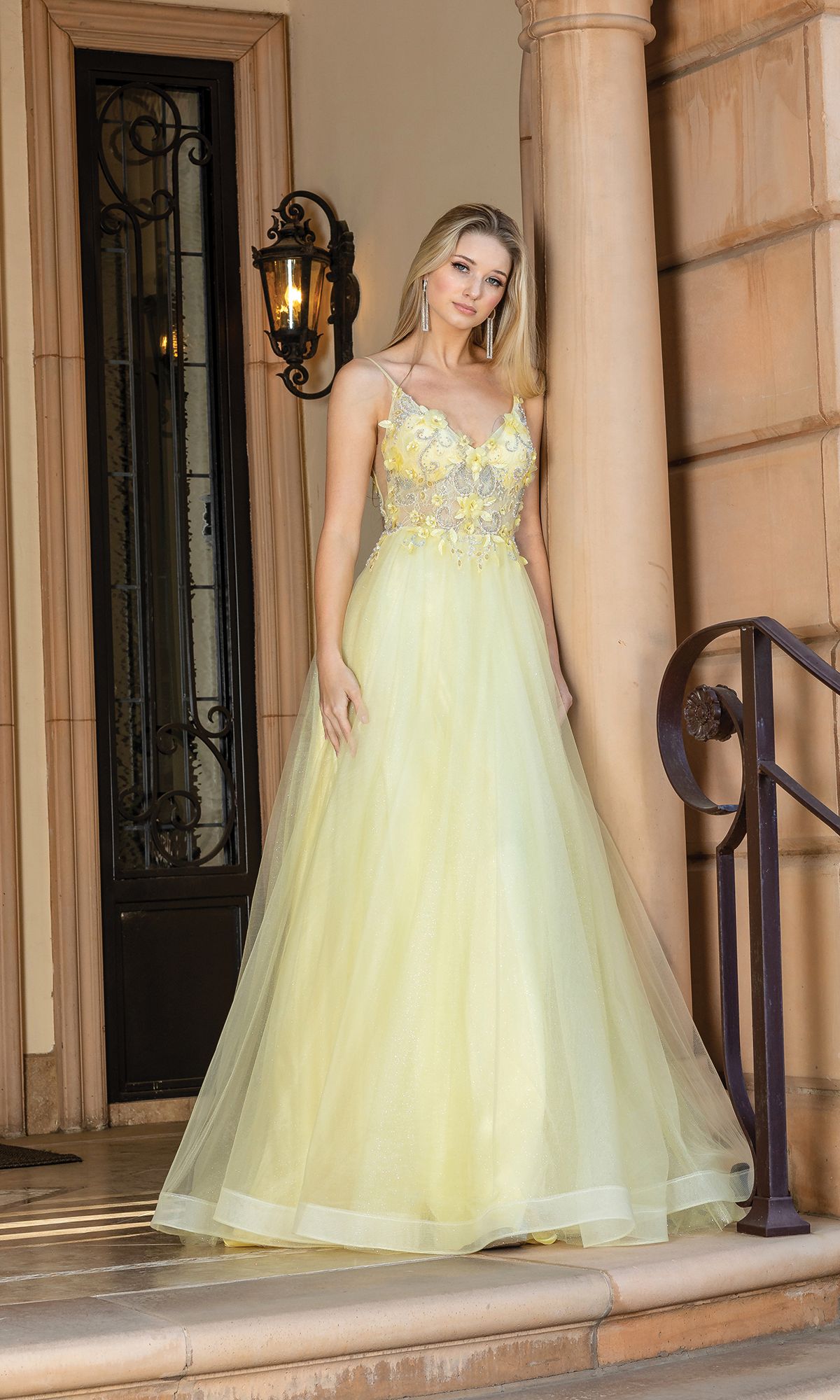 A woman stands on steps by a doorway, wearing the Long A-Line Tulle Prom Ball Gown with Flowers 4276—an elegant light yellow dress featuring 3-D floral designs and thin straps.