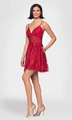A woman stands smiling against a plain background, wearing the Short Red Glitter Hoco Dress: Blondie Nites 4279BN with sequin details and spaghetti straps, paired with silver high-heeled shoes.