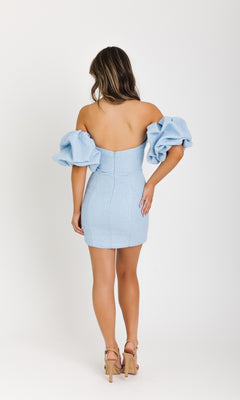 A woman faces away, wearing the Ava Presley 42913 short homecoming dress with off-the-shoulder ruffled sleeves and beige heels, standing against a plain white background.