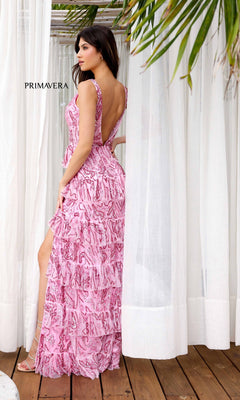 A woman wears the Long Formal Dress 4295 by Primavera, a pink ruffle-layered gown with a backless design, high slit, and elegant patterned details. She stands on a wooden floor near white curtains and palm leaves.