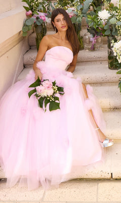 A person in a pink tulle Ava Presley 43019 strapless gown with 3D floral accents sits on stone steps holding a bouquet of pink and white flowers.