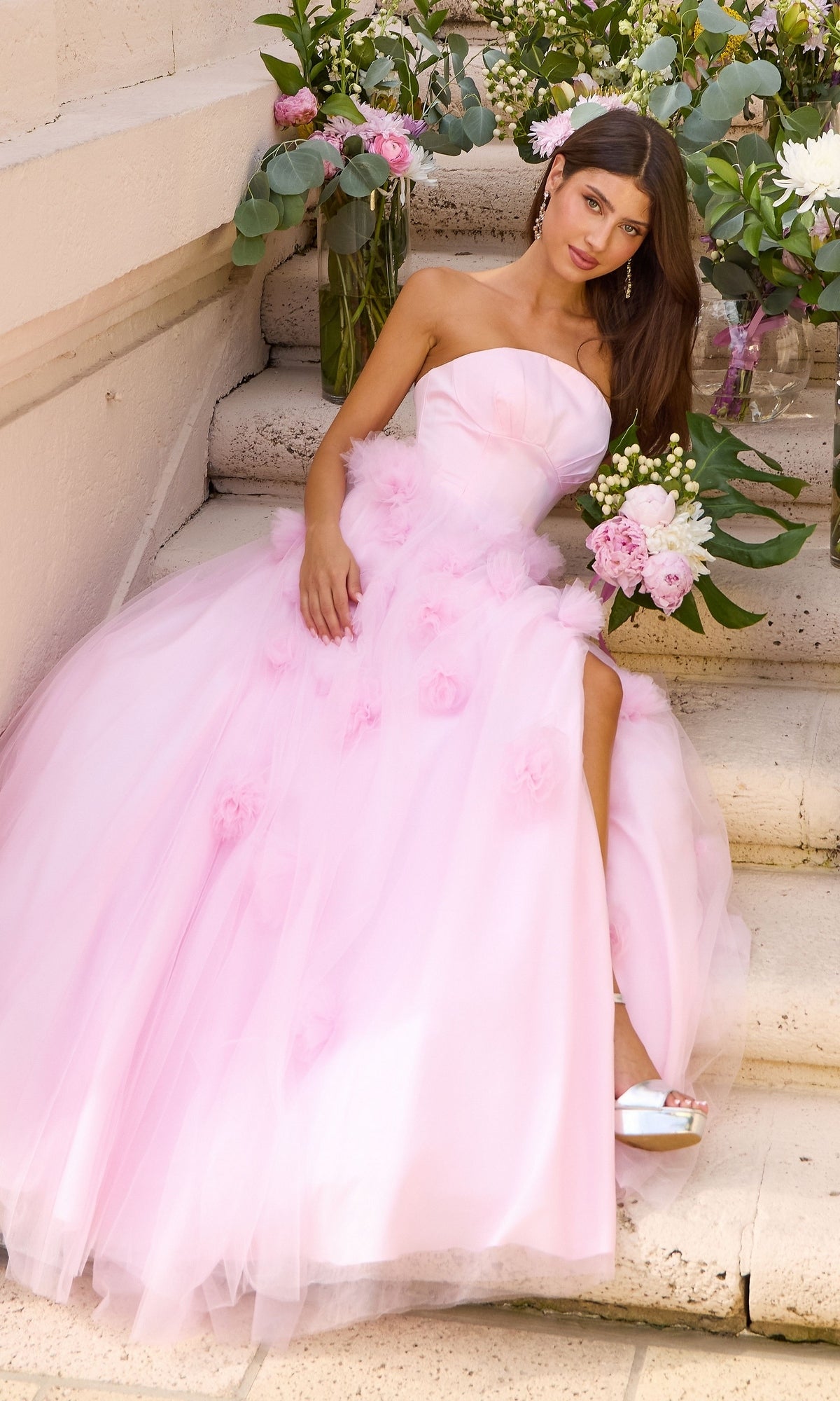 A person wearing a light pink, 3D floral, strapless Long Prom Dress: Ava Presley 43019 with a side slit, sitting on stone steps while holding a flower bouquet.