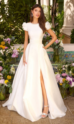 A woman in a white Ava Presley 43023 floor-length puff sleeve prom dress with a jewel neckline and high leg slit, standing among floral arrangements.
