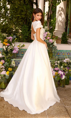 A person wearing a white, floor-length Ava Presley 43023 gown with puff sleeves and an open back, standing among arrangements of colorful flowers.