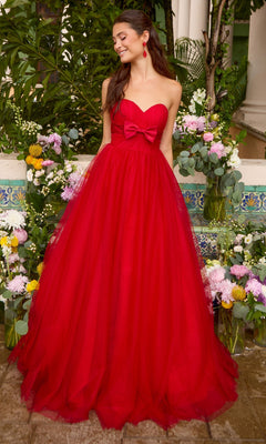 A person wears a red strapless floor-length Ava Presley 43045 ball gown with a sweetheart bodice and a bow, standing amidst floral arrangements.
