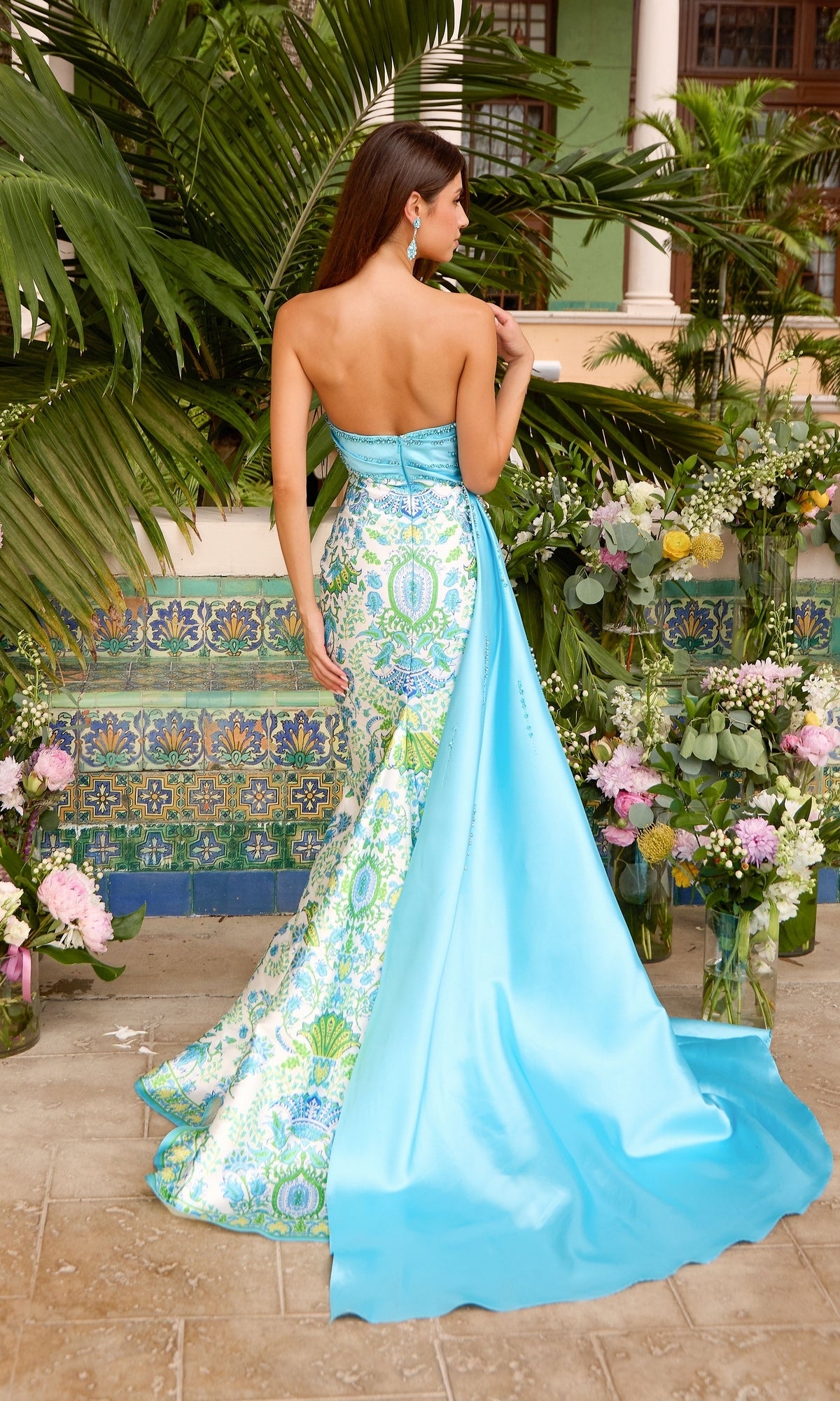 A woman wears an Ava Presley 43052 strapless, floor-length gown featuring a blue and white paisley print bodice and a solid blue skirt overlay with a train.