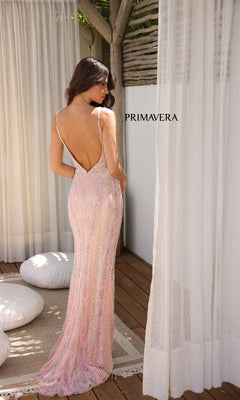 A woman stands on a wooden floor wearing the Primavera 4305, a fitted, light pink beaded long prom dress with adjustable spaghetti straps, backless design, and thigh-high slit. White curtains and cushions are behind her, with "PRIMAVERA" on the wall.