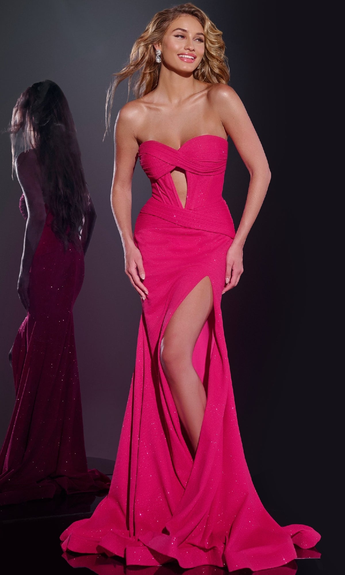 A woman wears the Jovani 43065 long prom dress, featuring a strapless glitter design and thigh-high slit, standing against a dark background with her reflection visible beside her.