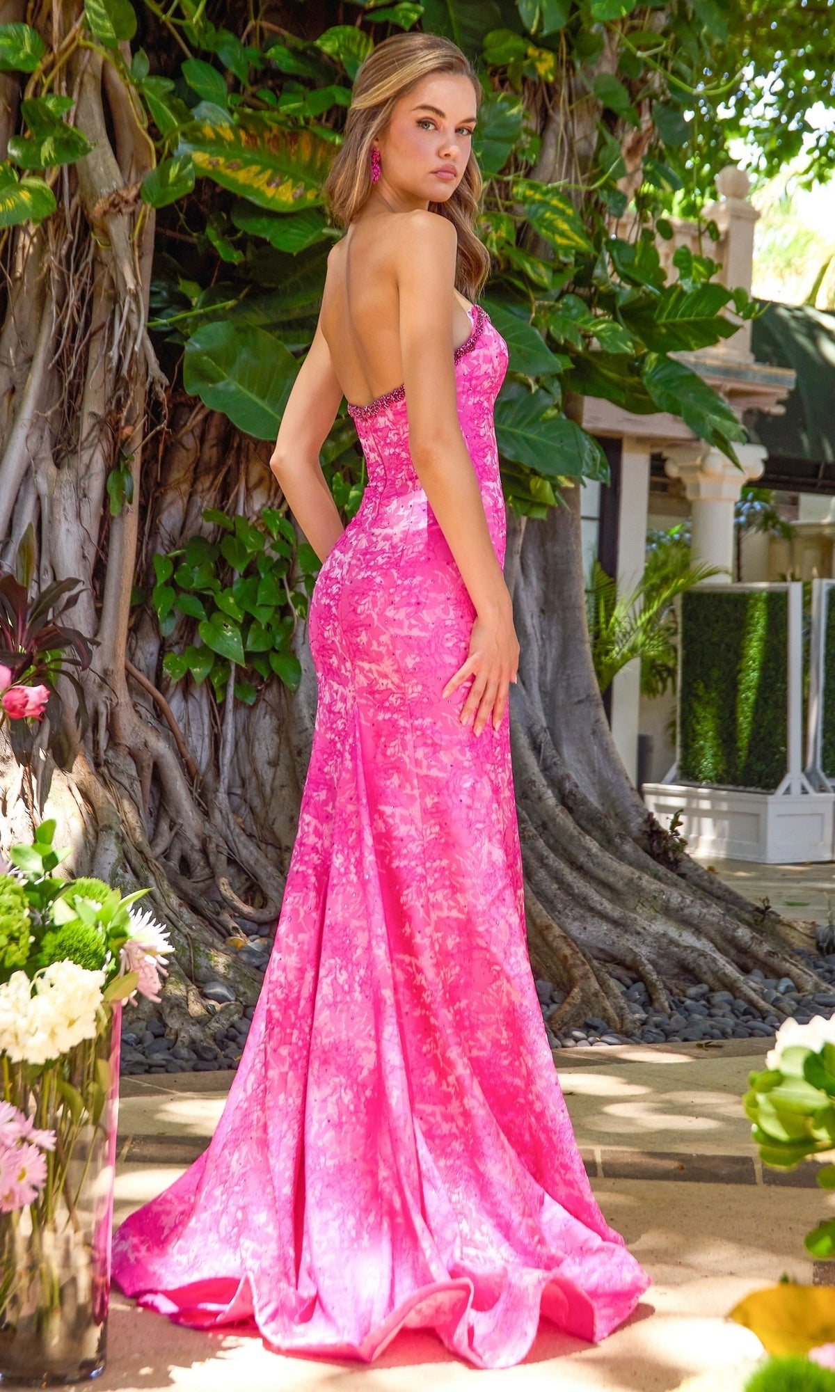 A woman wearing an Ava Presley 43077 floor-length, strapless bright pink mermaid prom dress with a beaded neckline, standing in front of a large tree.