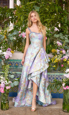 A person wears an Ava Presley 43090 light blue and purple floral brocade gown with a ruffled high-low hem and embellished straps, standing before a stone wall decorated with vases of flowers.