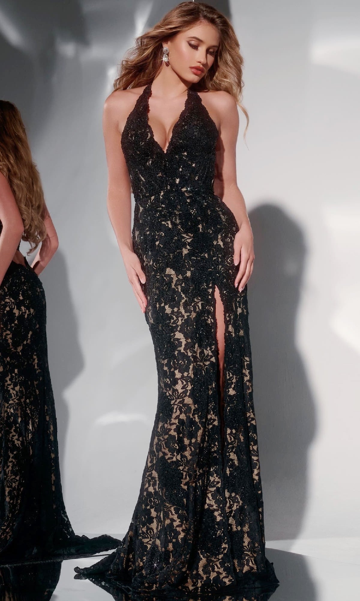 Woman wearing the Jovani 43097 long prom dress—a sleeveless black lace halter gown with a corset bodice, plunging neckline, and high slit—stands against a plain background.