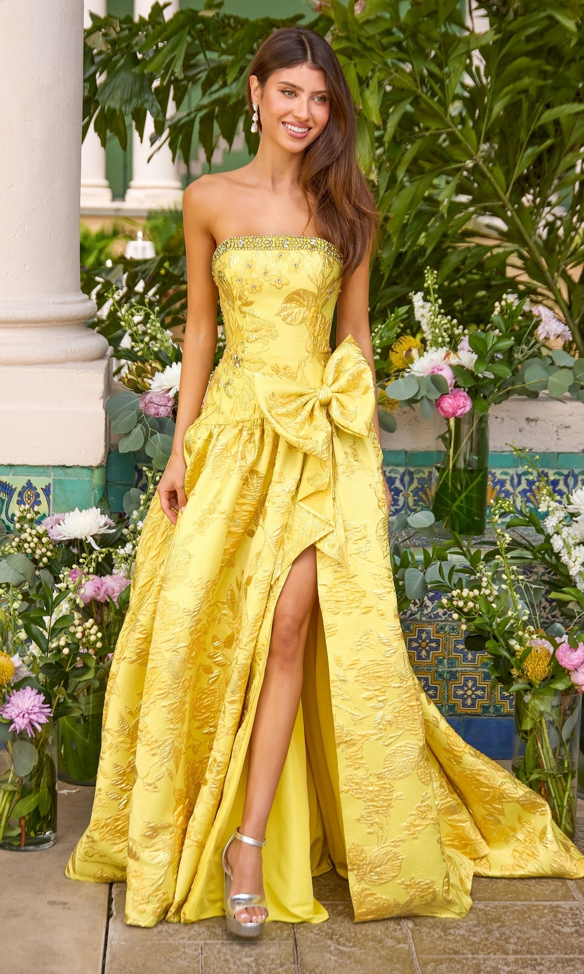 A strapless yellow Long Prom Dress: Ava Presley 43103 ball gown featuring a jacquard floral pattern, a large bow at the hip, and a high leg slit.