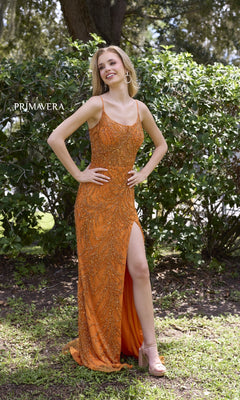 A woman poses outdoors in front of green bushes, wearing the Primavera 4310—a long beaded prom dress with a scoop neckline, sleeveless corset-style bodice, thigh-high slit, and vibrant orange hue.