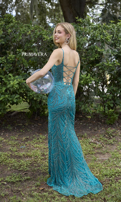 A woman stands on grass outdoors holding a clear confetti balloon, wearing the Primavera 4310 Scoop-Neck Long Beaded Prom Dress. Lush green bushes form the background.