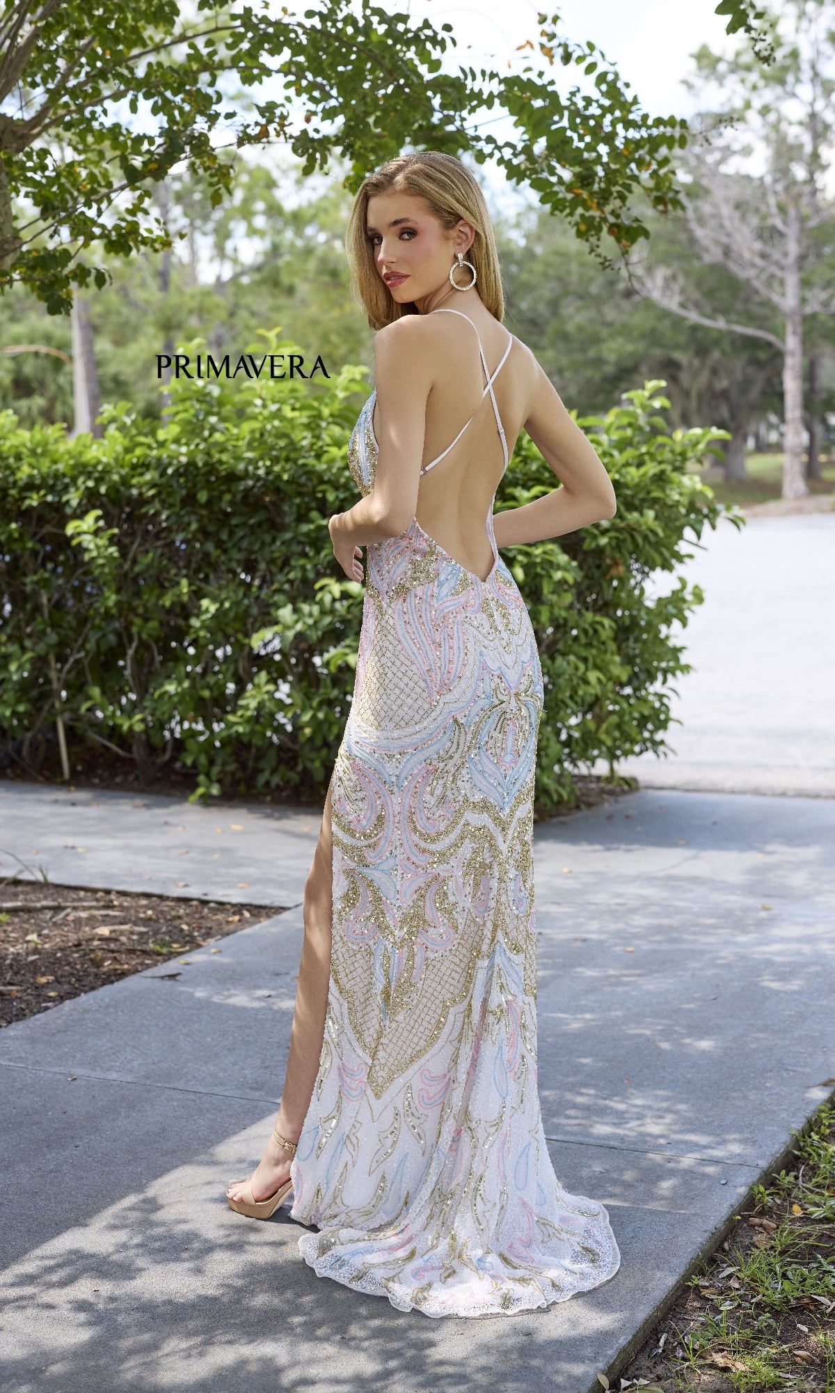 A woman wears the Primavera 4311, a long beaded V-neck prom dress with a backless design and side slit, standing outdoors on a sidewalk and looking over her shoulder.
