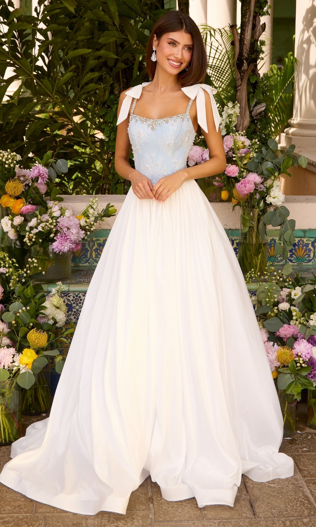 A woman wears a light blue and white Ava Presley 43128 long prom dress featuring an embellished bodice with tie-shoulder straps and a full skirt.