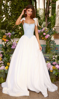 A woman wears an Ava Presley 43128 light blue prom dress with a beaded bodice, tie-shoulder straps, and a full white floor-length skirt while standing among arrangements of colorful flowers.