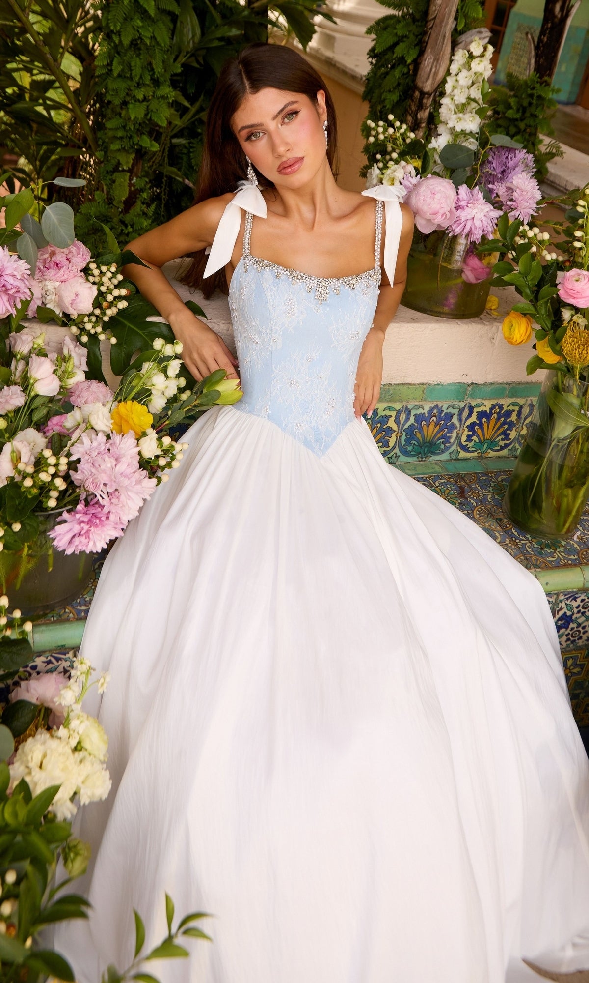 A woman wears a pale blue Ava Presley 43128 ball gown with a lace bodice, crystal-trimmed square neckline, white ribbon straps, and a full white skirt, surrounded by flower arrangements.