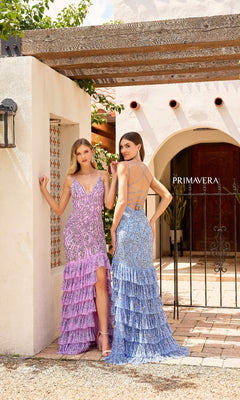 Two women pose outdoors in front of a white stucco building with arched doorways—one wears the Primavera 4315 purple glitter long prom dress, and the other wears a blue gown with a ruffled skirt.