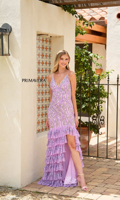 A woman stands outdoors in front of a white wall, wearing the Primavera 4315 Long Prom Dress featuring lilac sequins, a sleeveless design, ruffled skirt, and high-low hem, paired with open-toed heels.